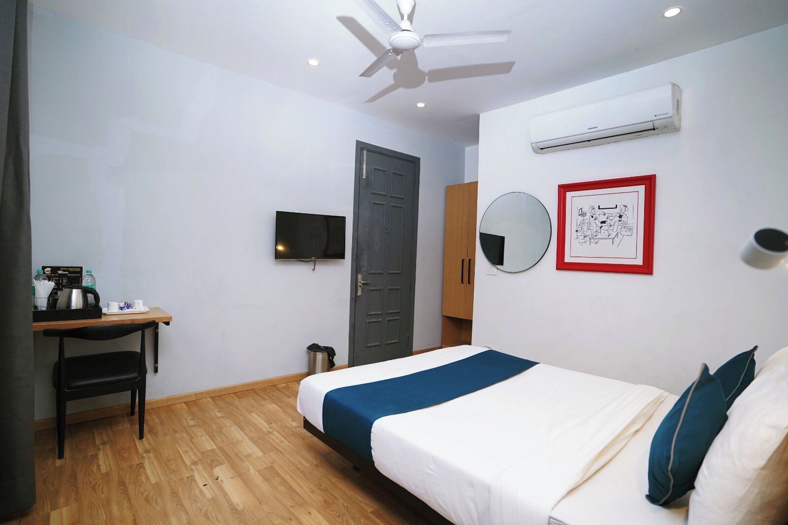 A hotel room with a bed, a desk, a chair, a television, a wardrobe, a mirror, and an air conditioning unit.