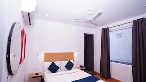 Quarto de hotel com cama, mesa de cabeceira, ar condicionado fixado na parede, ventilador de teto, janela com cortinas e uma porta.