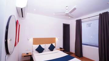A hotel room with a bed, bedside table, nightstand, wall-mounted air conditioner, ceiling fan, window with curtains, and a door.