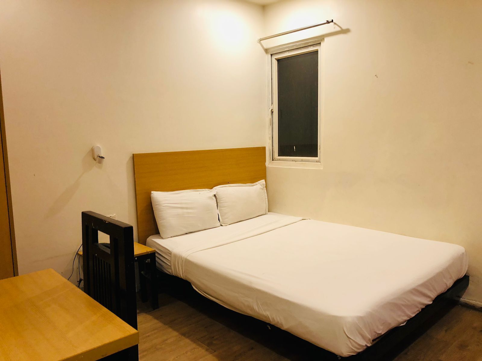A single bed with white bedding, a wooden headboard, a small bedside table, and a window with a blind.