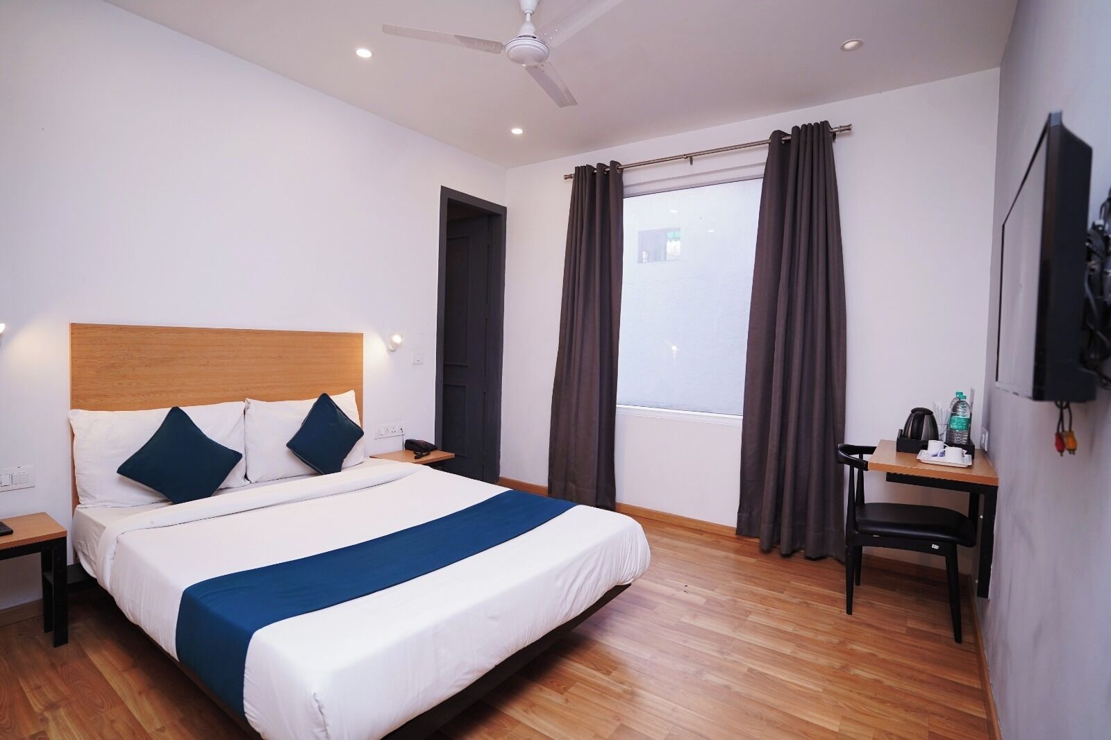 A hotel room with a bed, a desk, a chair, a window with curtains, a ceiling fan, and a wall-mounted TV.
