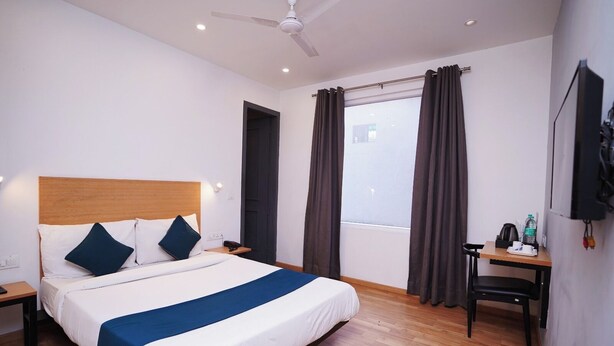 Quarto de hotel com cama, escrivaninha, cadeira, janela com cortinas, ventoinha no teto e televisão presa na parede.