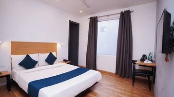 A hotel room with a bed, a desk, a chair, a window with curtains, a ceiling fan, and a wall-mounted TV.