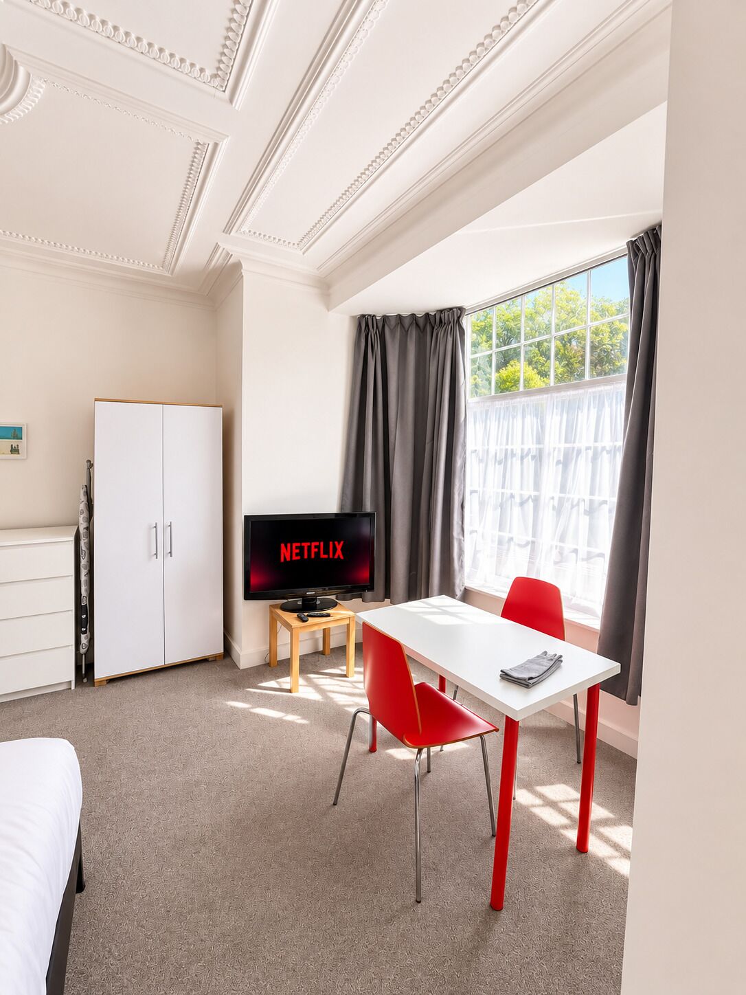 A hotel room with a white wardrobe, a television displaying 'NETFLIX', a white table with a red chair, and a window with curtains.