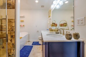 A modern bathroom with a stone wall, a large mirror, a white sink, a gold-toned vanity, and a blue bath mat. - Home Gym & Pool Access! North Raleigh Retreat (Raleigh)