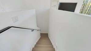 Un escalier étroit aux murs blancs et au plancher en bois, menant à une pièce où se trouve un téléviseur de couleur sombre.