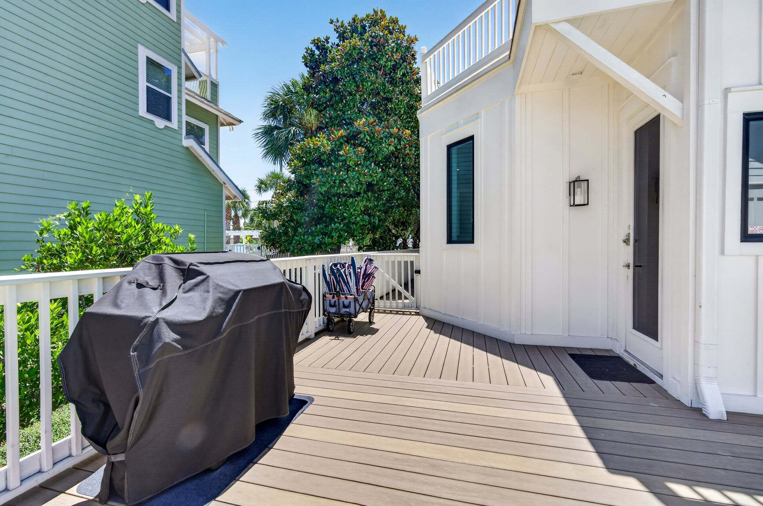 A covered outdoor deck with a barbecue grill and a cart of items, adjacent to a white building with a door and a window.