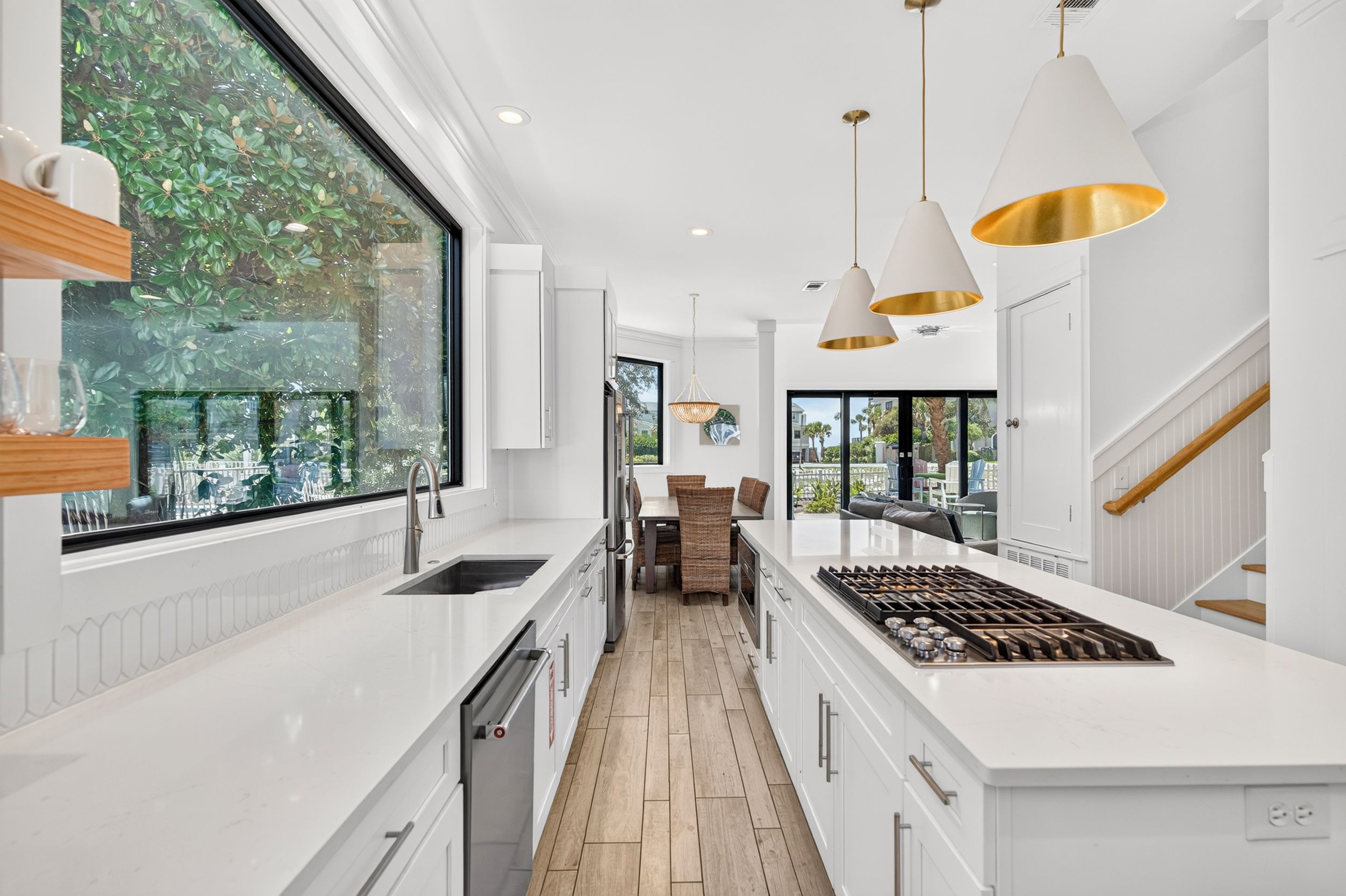 Una cocina moderna con armarios blancos, una isla central con estufa, iluminación colgante y un amplio ventanal con vistas a zonas verdes.