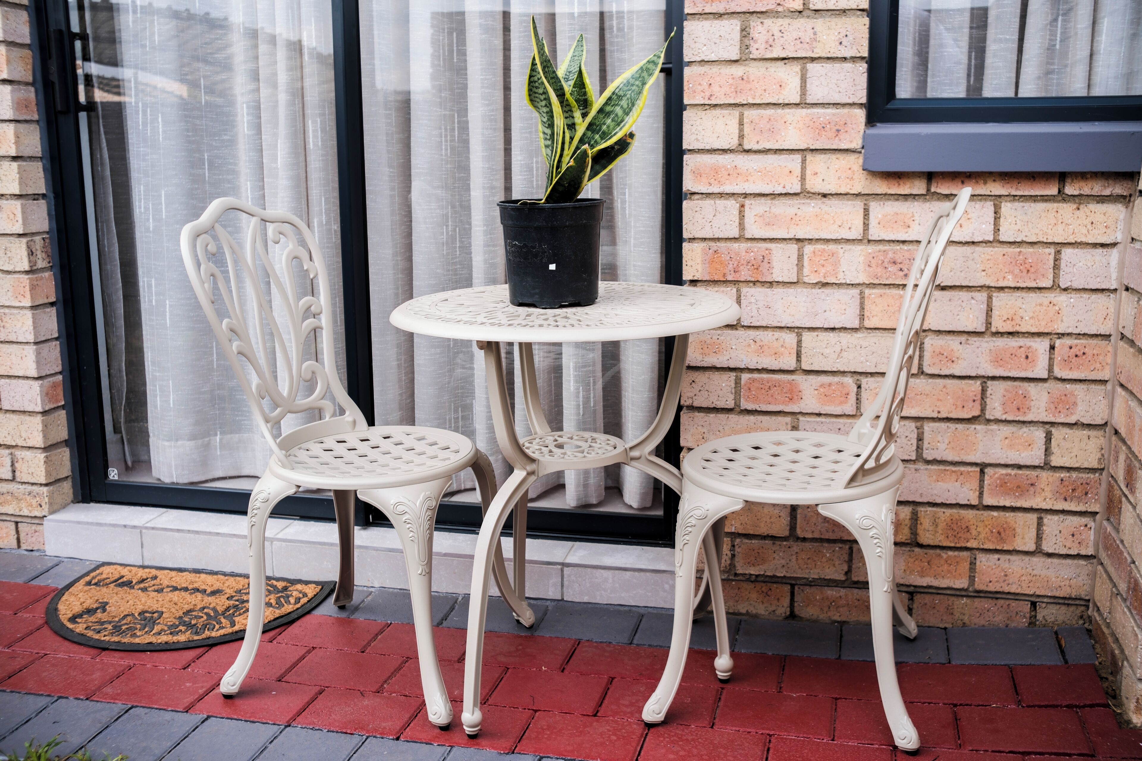 A patio with two white metal chairs and a small round table, a potted plant, and a doormat.