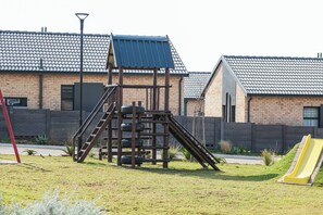 A playground with a slide and climbing structure, surrounded by a grassy area and residential buildings in the background. - Cozy 1-bedroom Apartment in Secure Estate, Tshwane (Centurion)