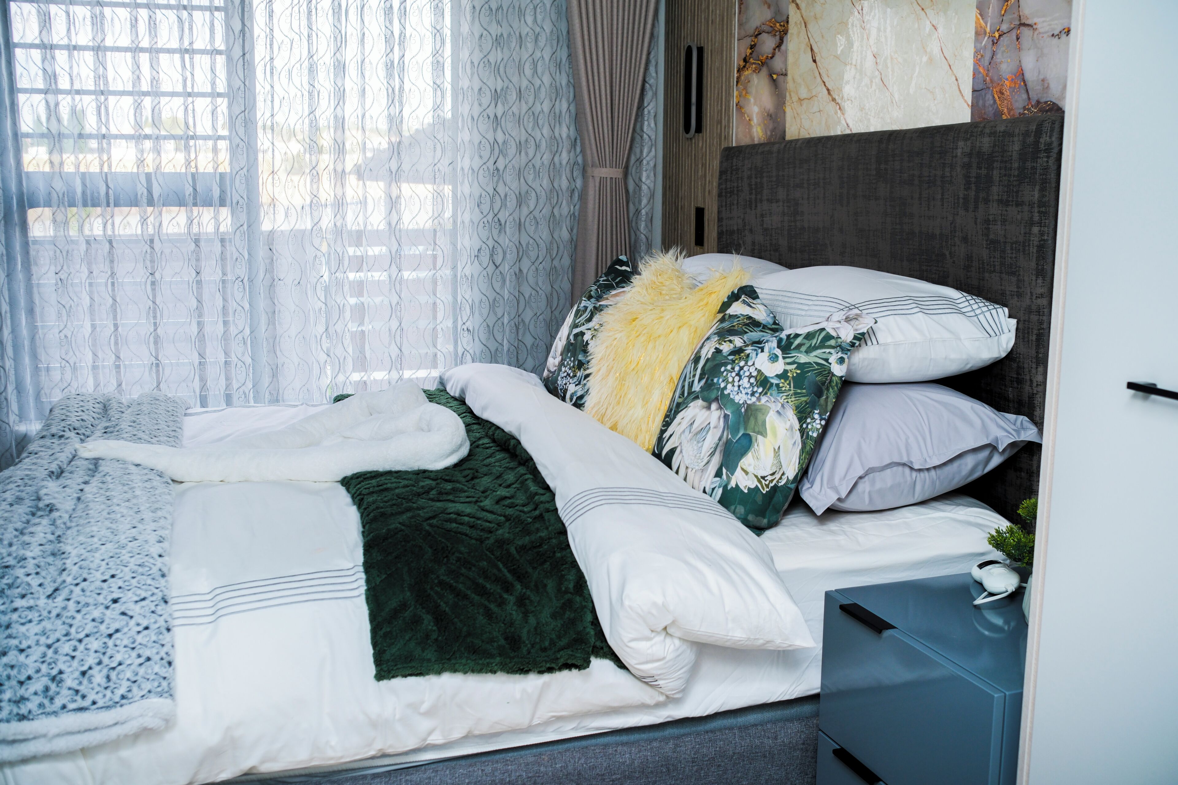 A neatly made bed with a variety of pillows and a blanket, a bedside table with a lamp, and a view of the outside through sheer curtains.