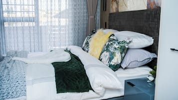 A neatly made bed with a variety of pillows and a blanket, a bedside table with a lamp, and a view of the outside through sheer curtains.