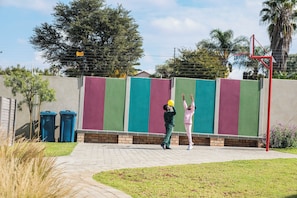 A child playing with a ball near a basketball hoop, with a colorful fence and trash bins in the background. - Cozy 1-bedroom Apartment in Secure Estate, Tshwane (Centurion)