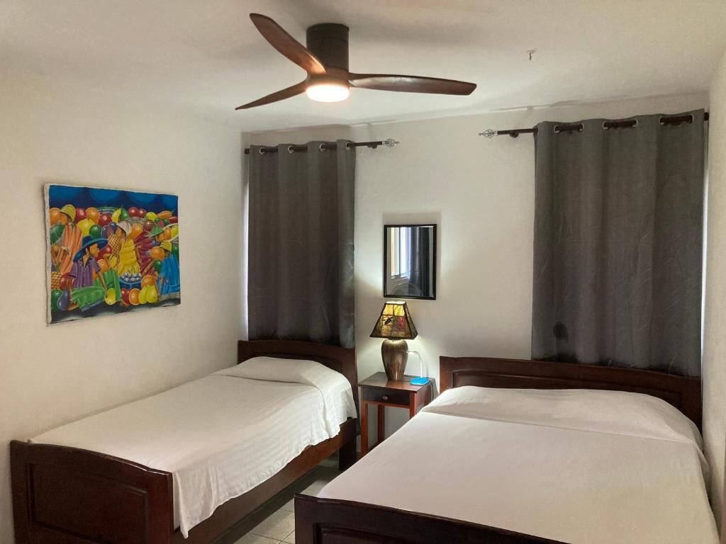 Ein Hotelzimmer mit zwei Einzelbetten, einem Deckenventilator, einem Bild an der Wand, einer Lampe und einem Fenster mit Vorhängen.
