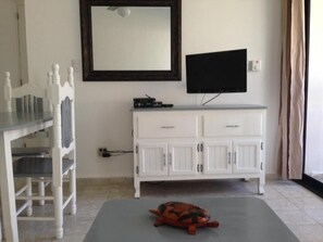 A room with a white cabinet, a television mounted above it, a small turtle on a mat, and a chair with a table. - Charming 1-bedroom Apartment in Vibrant Cabarete, DR (Sosúa)