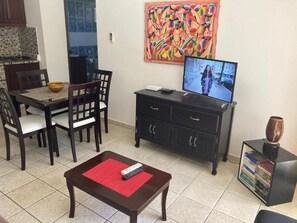 A living room with a dining table, chairs, a TV, a coffee table, and a bookshelf. - Charming 1-bedroom Apartment in Vibrant Cabarete, DR (Sosúa)