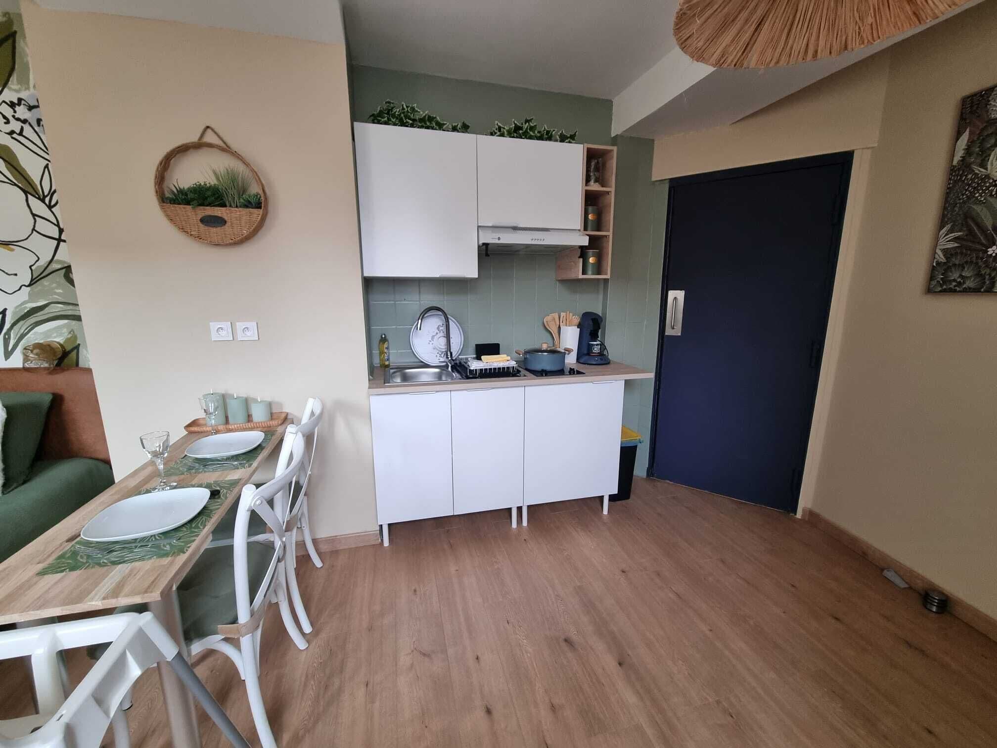 Una cocina compacta con armarios blancos, una mesa de comedor con platos y cubiertos, y una pequeña isla con una planta.