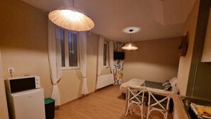 A compact living space with a kitchenette, a dining area with a table and chairs, and a ceiling light fixture. - Le Pti Marais, a 3-star studio on the outskirts of Saint-Omer (Saint-Martin-lez-Tatinghem)