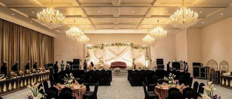 Une salle de banquet avec des tables rondes, des chaises noires et une scène décorée de fleurs.