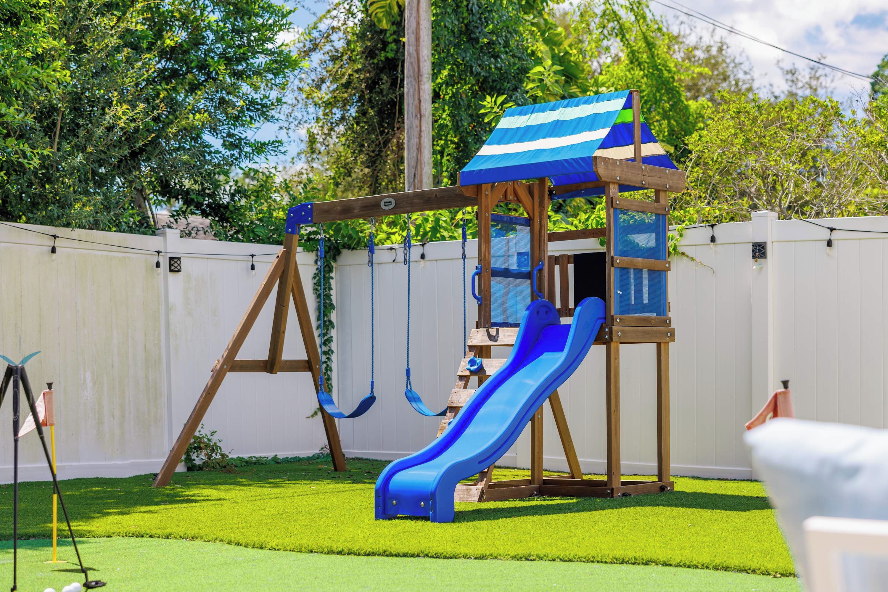 A wooden play structure with a slide and swings, set against a white fence and green grass.