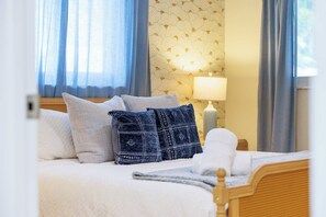 Cama bem arrumada com roupa de cama branca e alguns travesseiros, uma luminária de cabeceira e uma janela com cortinas.