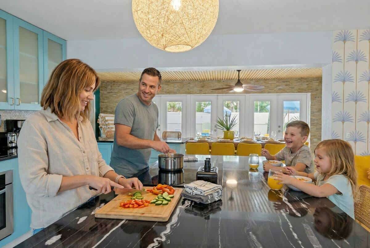 Eine Familie bereitet Essen in einer modernen Küche mit einer großen Kochinsel zu, mit einem Essbereich mit gelben Stühlen und Blick ins Freie.