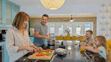 Eine Familie bereitet Essen in einer modernen Küche mit einer großen Kochinsel zu, mit einem Essbereich mit gelben Stühlen und Blick ins Freie.