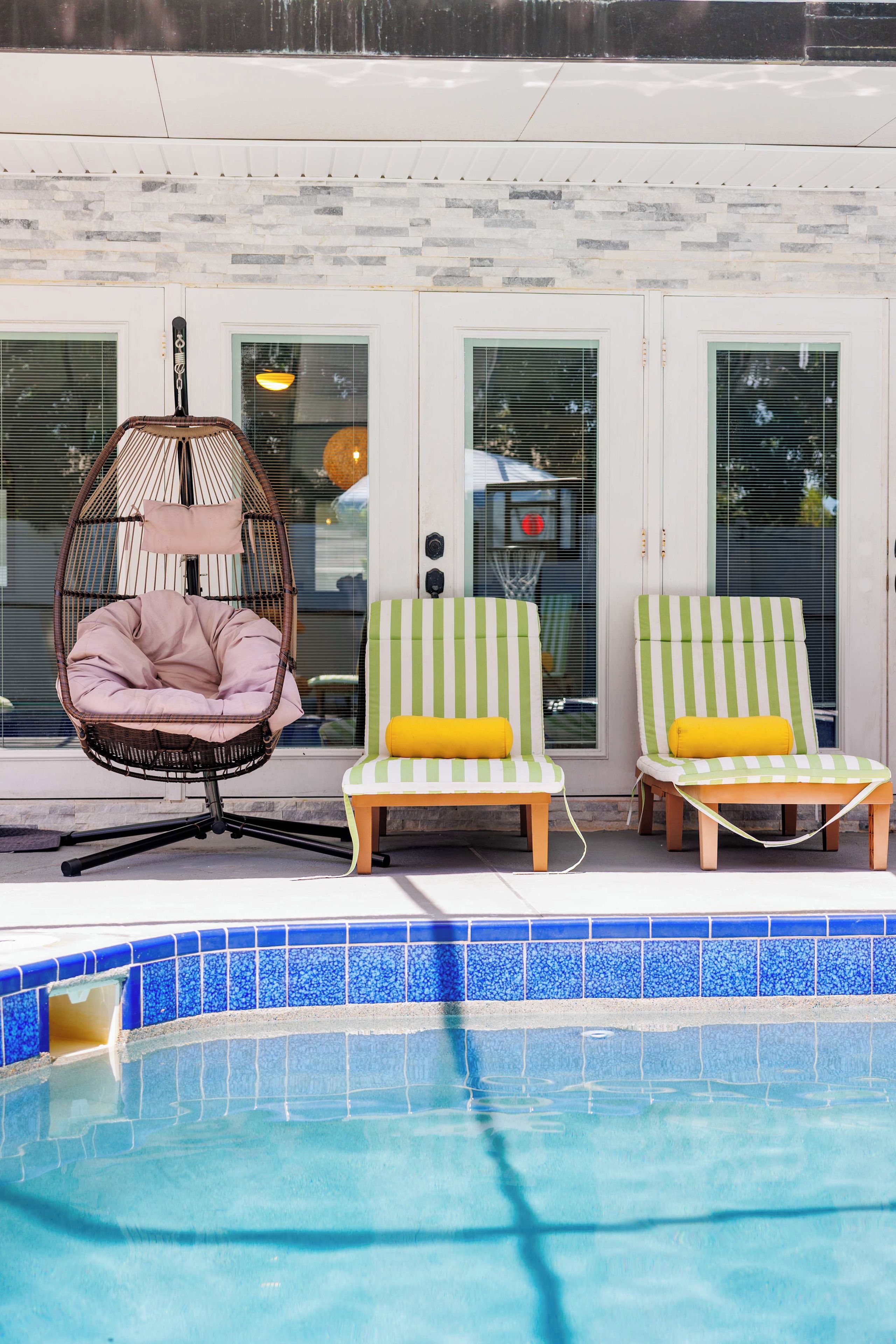 A poolside area with a hanging chair, two striped lounge chairs, and a pool with blue tiles.
