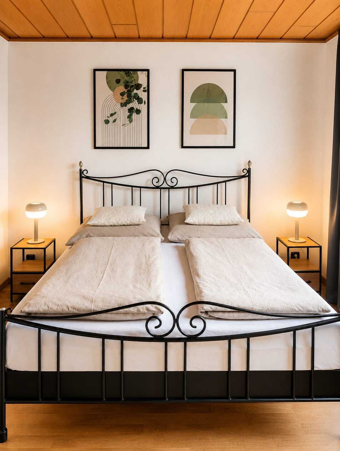 A bed with a black metal frame, two bedside tables with lamps, and two framed artworks on the wall.