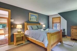 A bedroom with a wooden bed, two bedside tables with lamps, a framed picture on the wall, and a view through a window with blinds. - Deck & Wooded Views! 2 Mi to Frederick Gunn School (Washington)