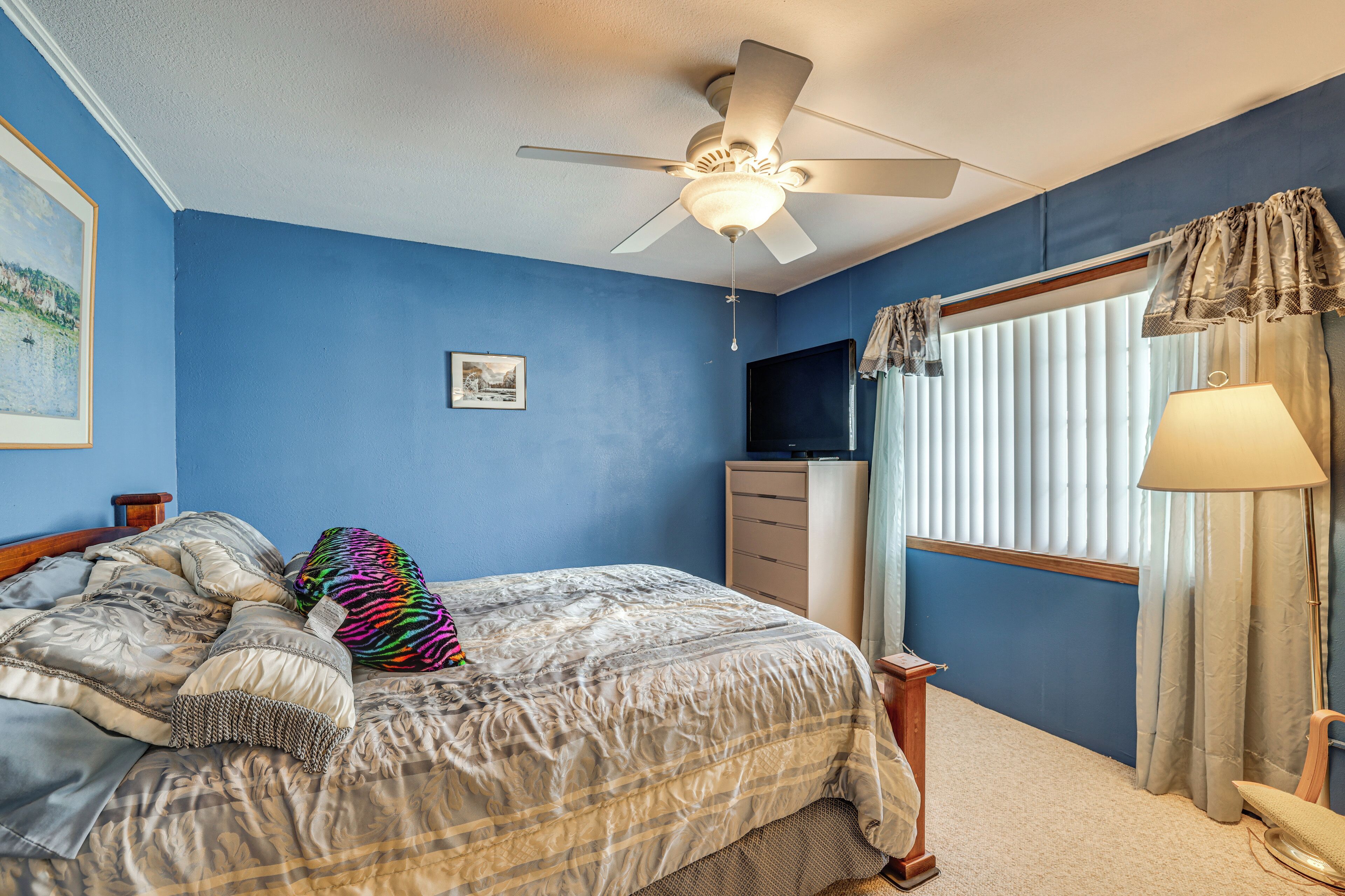 A bedroom with a bed, a ceiling fan, a dresser, a window with curtains, and a lamp.