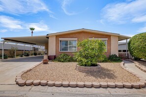 A single-story house with a flat roof, a front porch, and a small garden area with a variety of plants and a gravel driveway. - Palm Desert Home in 55+ Golf Community w/ Pool (Palm Desert)
