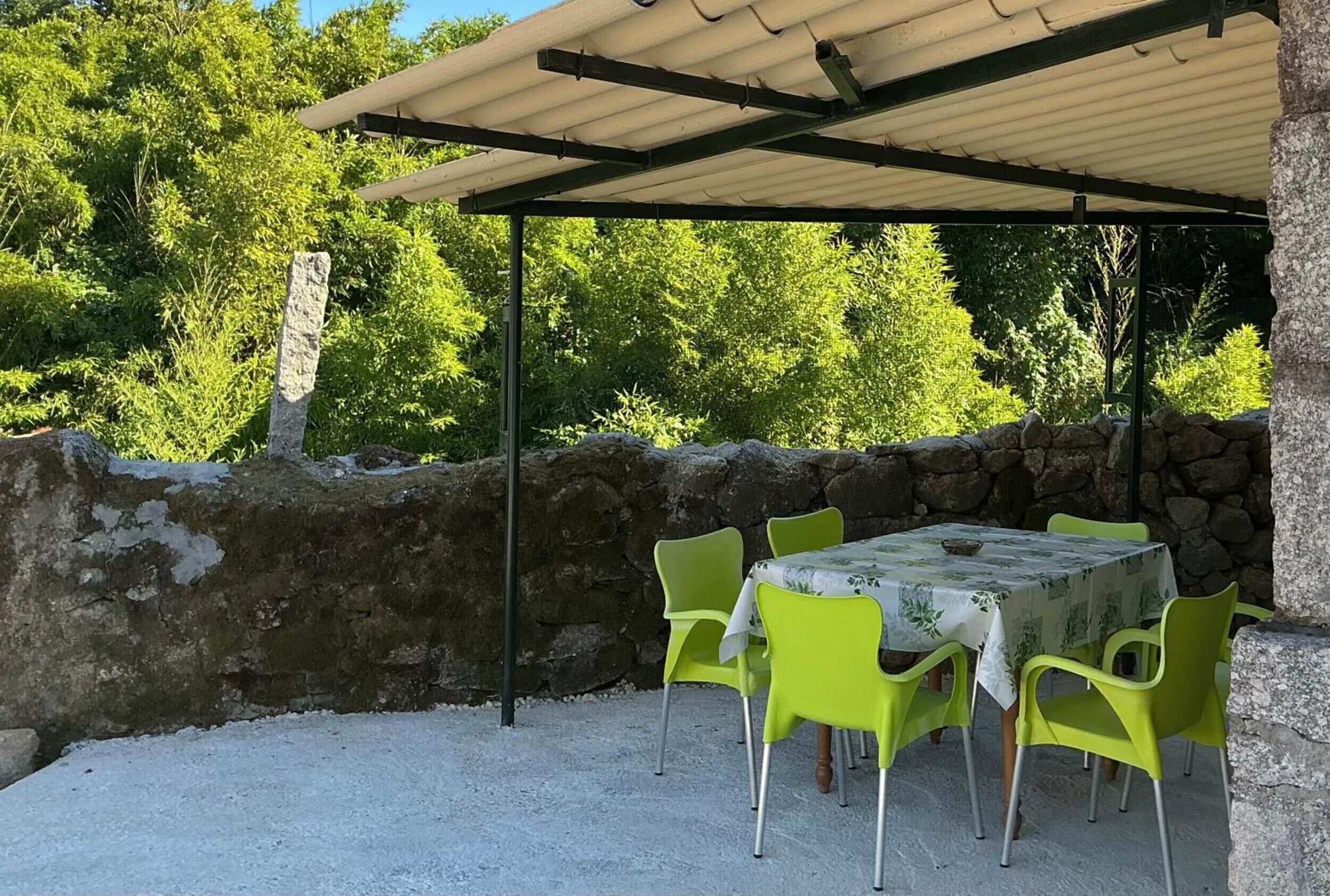 En udendørs terrasse med et bord, stole, en stenvæg og et overdækket område med et tag.