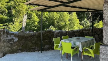 En udendørs terrasse med et bord, stole, en stenvæg og et overdækket område med et tag.