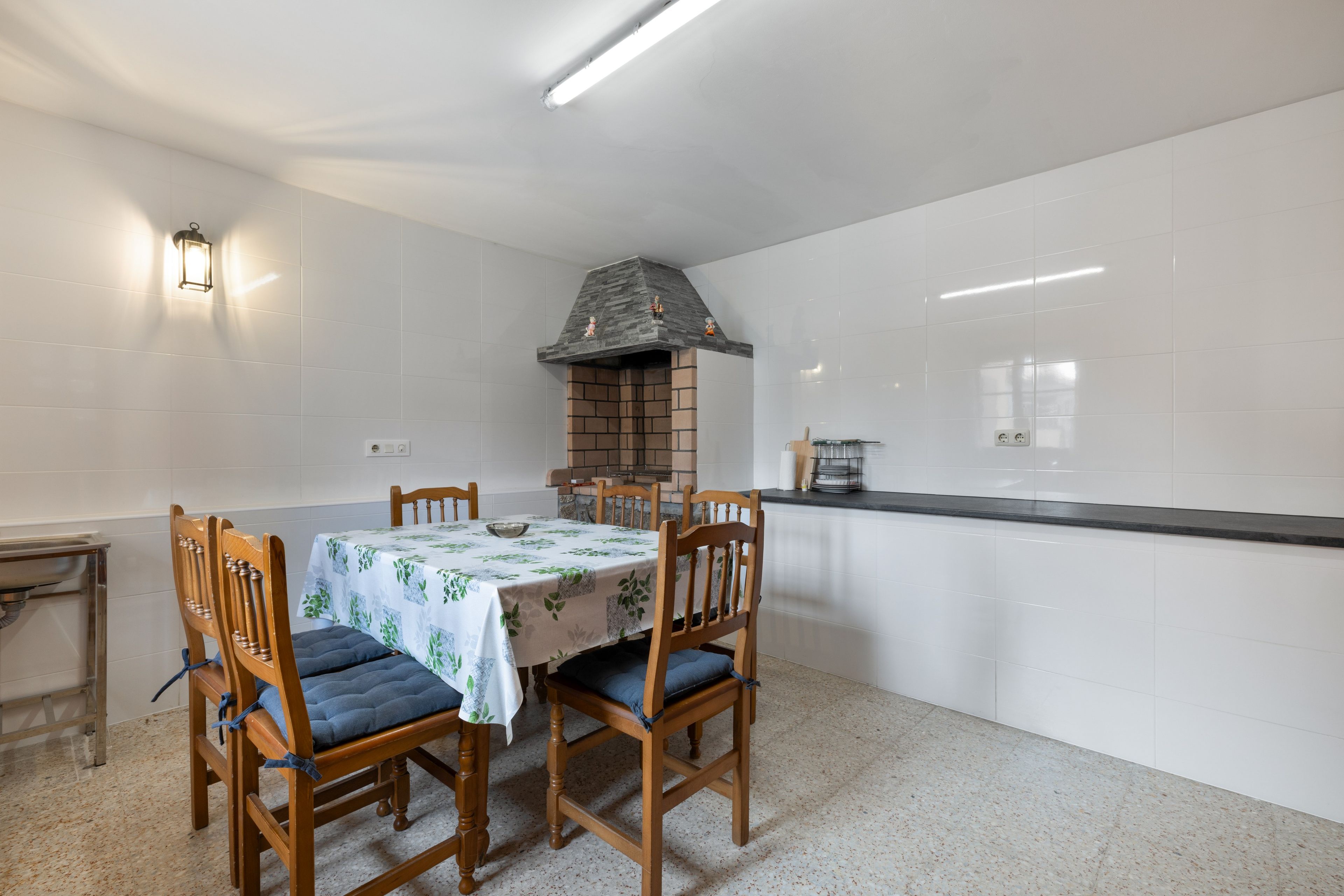 Un comedor con una mesa dispuesta para cuatro personas, rodeada de sillas de madera con cojines azules. Un hogar de ladrillo con chimenea se encuentra centralmente sobre la mesa.