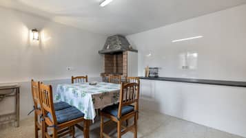 Un comedor con una mesa dispuesta para cuatro personas, rodeada de sillas de madera con cojines azules. Un hogar de ladrillo con chimenea se encuentra centralmente sobre la mesa.
