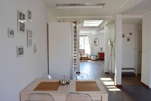 A dining area with a table set for two, a chair, and a view into a living room with a sofa and a window. - Rooftop terrace loft apartment, home cinema (Praha 10)