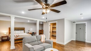A spacious room with a ceiling fan, a bed with a wooden headboard, a grey sofa, a small round table, and a bathroom visible through an open door.