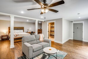 A spacious room with a ceiling fan, a bed with a wooden headboard, a grey sofa, a small round table, and a bathroom visible through an open door.