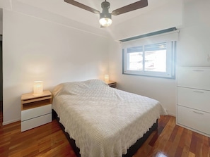 Un dormitorio con una cama, ventilador de techo, ventana y mesita de noche con lámpara.