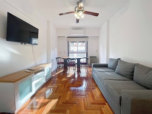 Una sala de estar moderna con un sofá gris, suelo de madera en espiga, ventilador de techo y televisor montado en la pared.