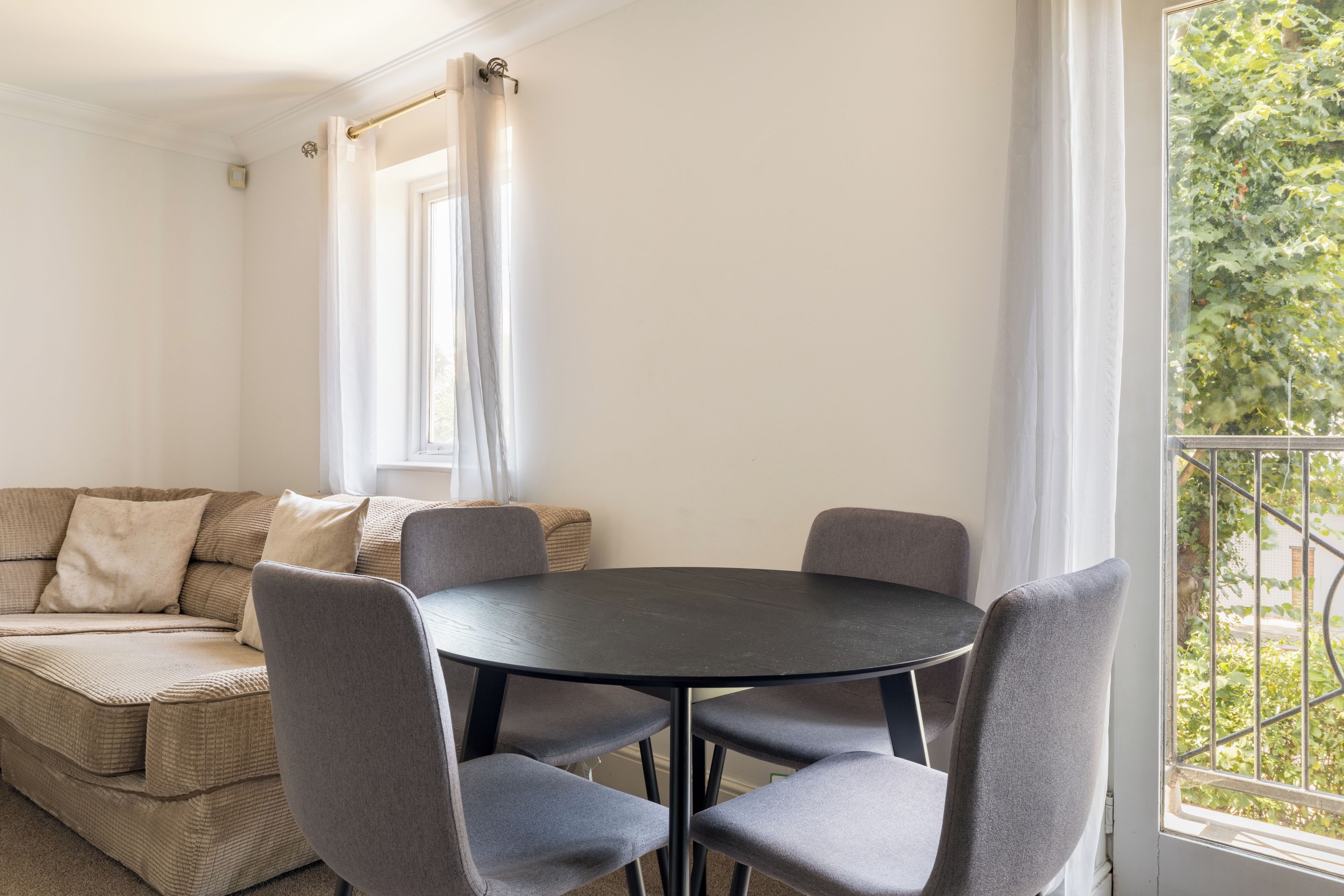 A room with a round table and four chairs, a sofa with cushions, a window with curtains, and a view of greenery outside.