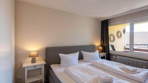 Quarto de hotel com cama, mesas de cabeceira, janela com cortinas e decorações nas paredes.