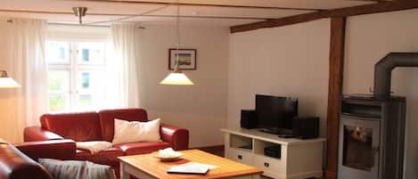 Un salon avec un canapé en cuir rouge, une table basse en bois, un téléviseur posé sur un meuble blanc et un poêle à bois.