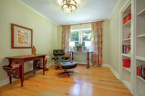 A room with hardwood flooring, a black leather chair, a wooden table with a bottle, a framed picture, a window with curtains, and a bookshelf.
