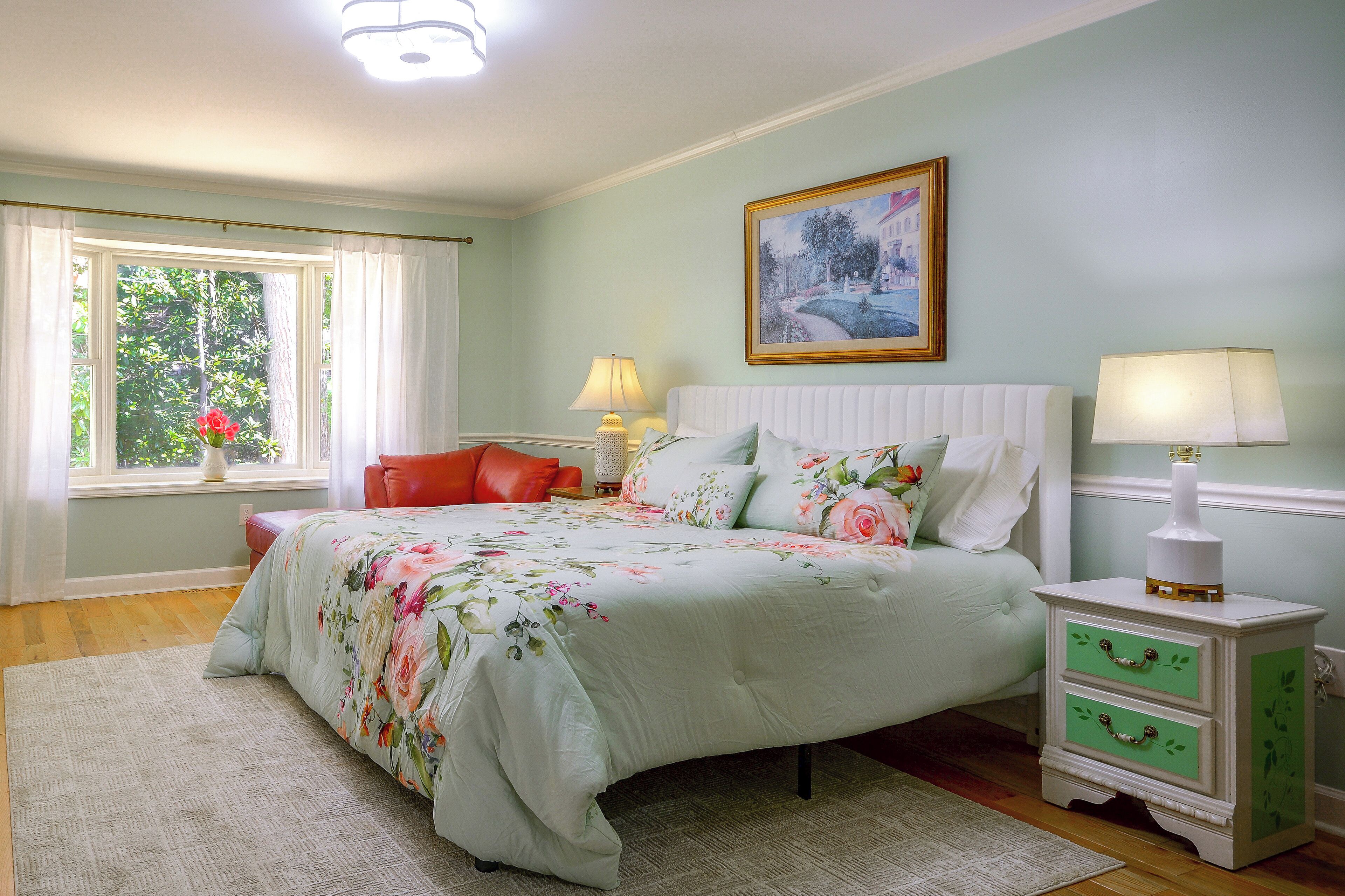 A bedroom with a bed, bedside tables, a window with curtains, a painting on the wall, and a view of greenery outside.
