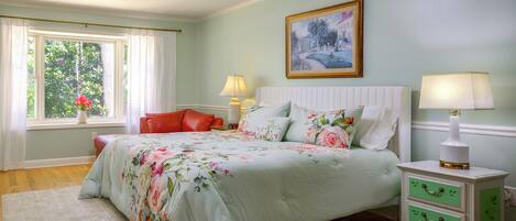A bedroom with a bed, bedside tables, a window with curtains, a painting on the wall, and a view of greenery outside.