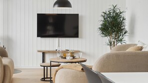 Moderni olohuone, jossa seinälle on asennettu taulutelevisio, huoneessa on pyöreä puinen pöytä, jonka päällä on maljakko ja laseja, beige sohva ja ruukkukasvi.