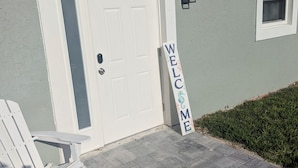 Una casa con una porta bianca, un cartello di benvenuto, una panchina e una piccola finestra.