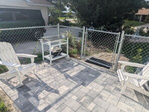 A patio area with a grill, two Adirondack chairs, and a chain-link fence. - Cozy Coastal Suite  (Bradenton)